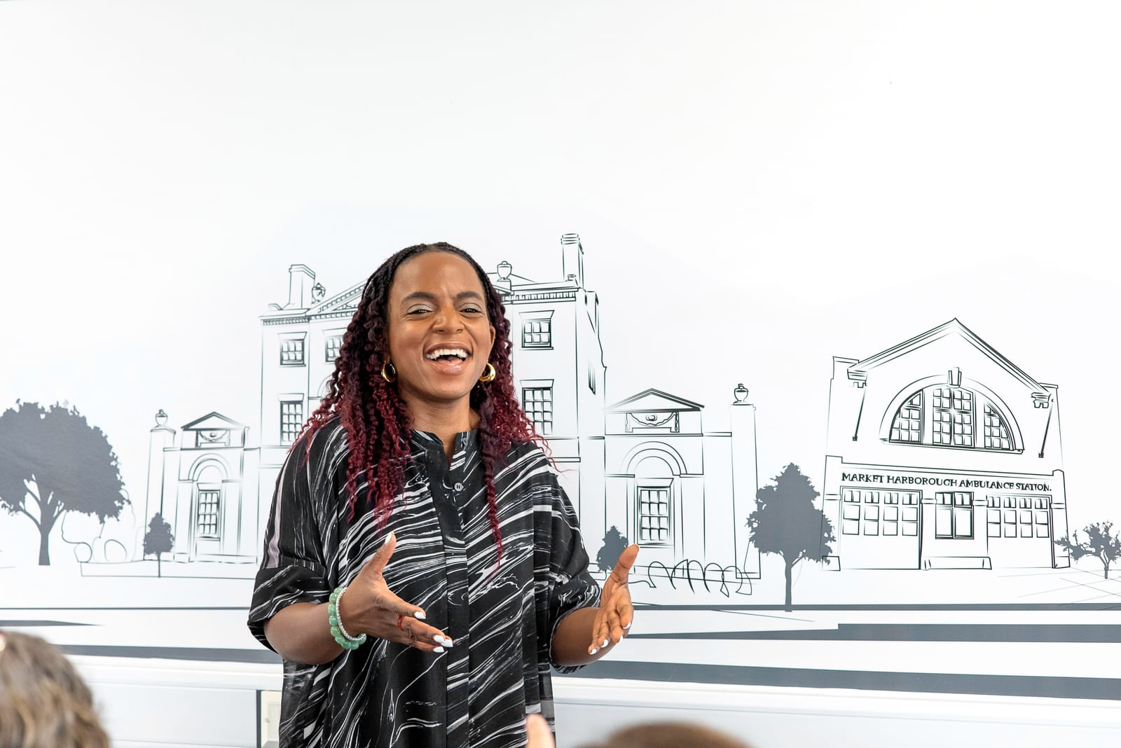 Blended Learning Studio facilitator laughing with arms open in front of a Market Harborough line-art mural while delivering a session.